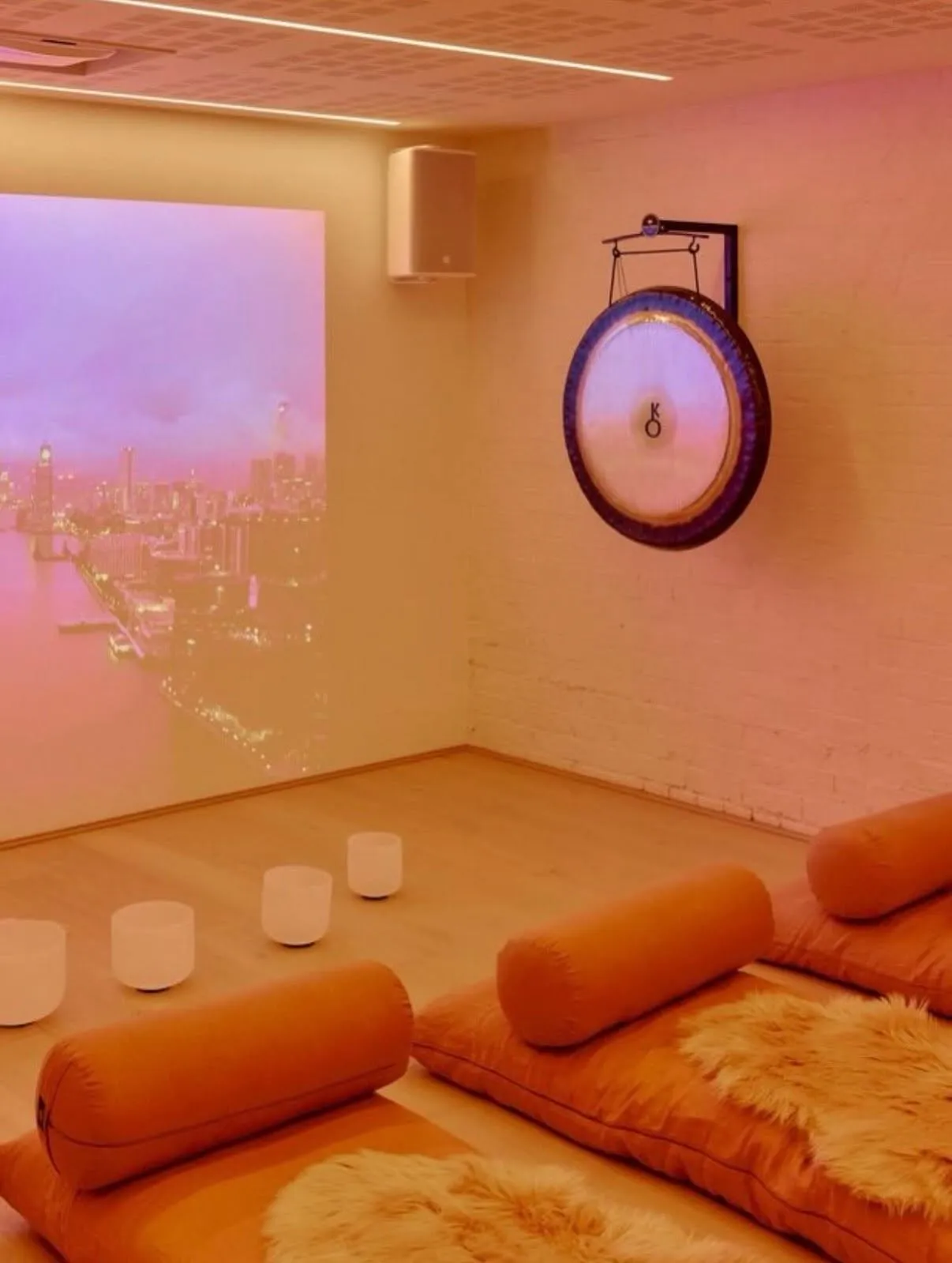 The downstairs studio in a warm pink light: a gong on the brick wall, crystal singing bowls and floor mats with sheepskins